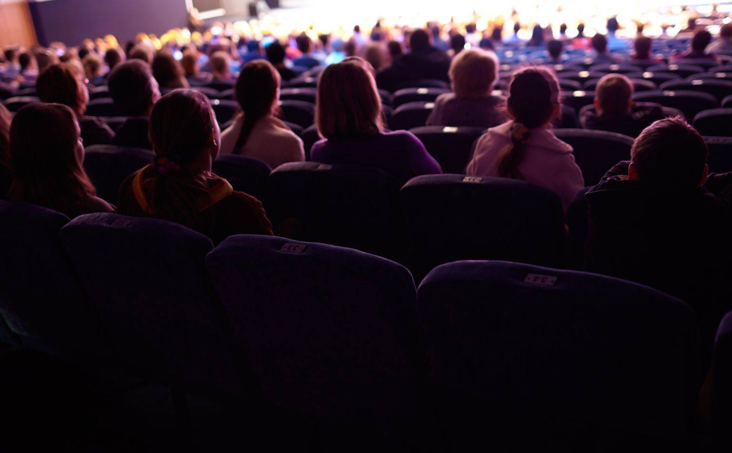 Widownia w kinie widziana od tyłu, na ekranie koncert w ciepłym świetle scenicznym